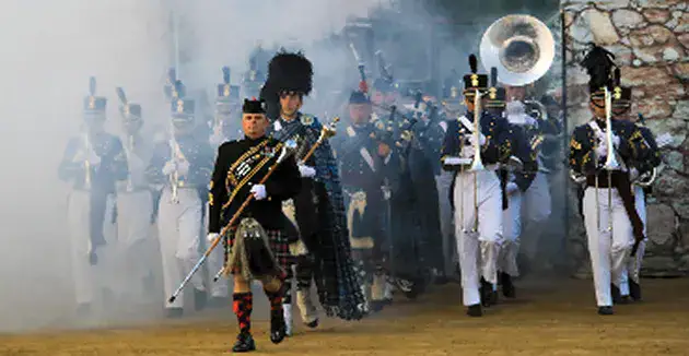 A drum major leading pipers and ceremonial musicians through smoke during an outdoor performance.